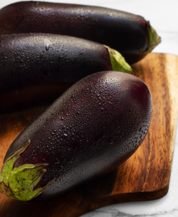 Tres berenjenas moradas con gotas de agua sobre una tabla de cortar de madera.