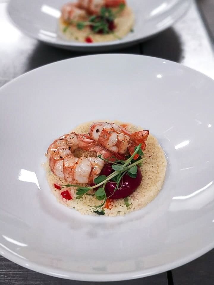 Antipasto di gamberi con erbe aromatiche e salsa servito in una ciotola bianca, con un altro piatto sullo sfondo sfocato.