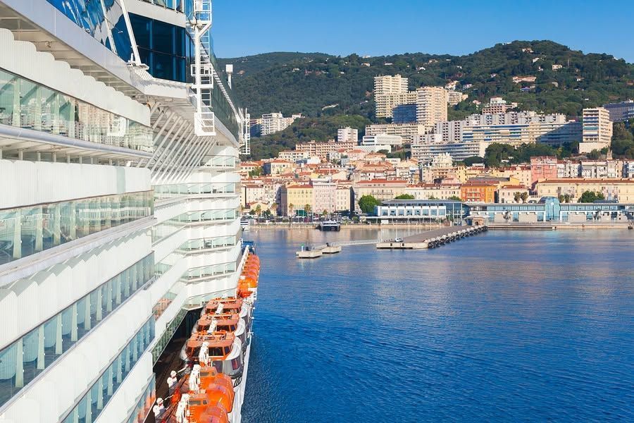 Cruise ship docked in a sunny harbor with blue water and hillside buildings in the background