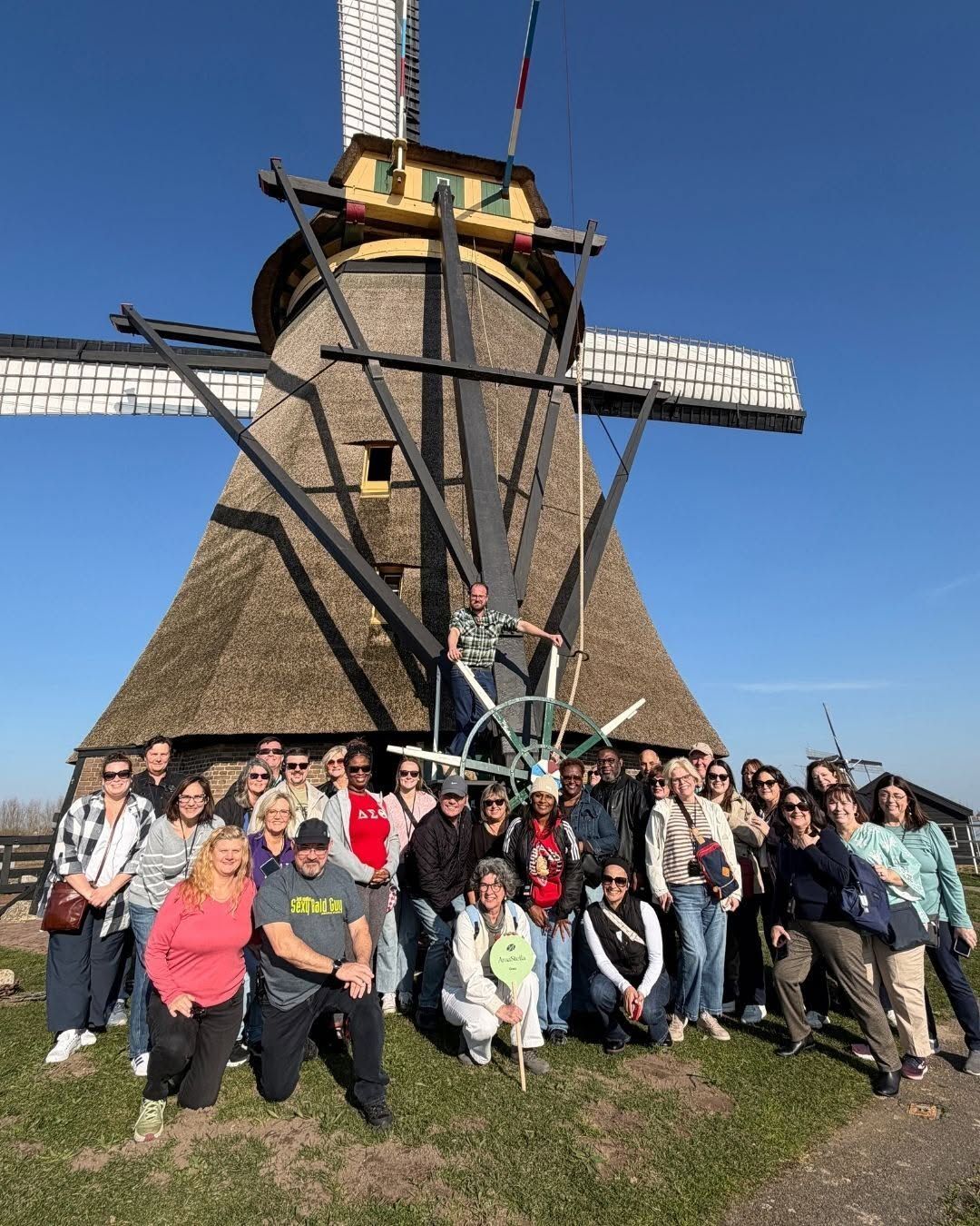 Group posing in front of a large windmill on a sunny day, with blue sky and grass foreground