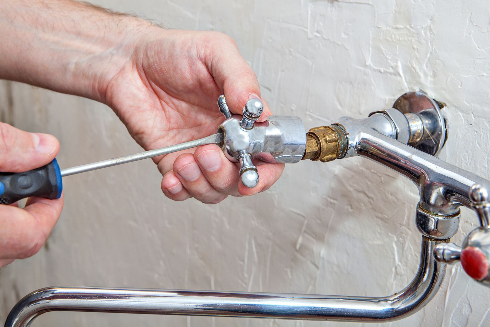 Close-up of hands using a screwdriver to fix water faucet handle on a white wall.