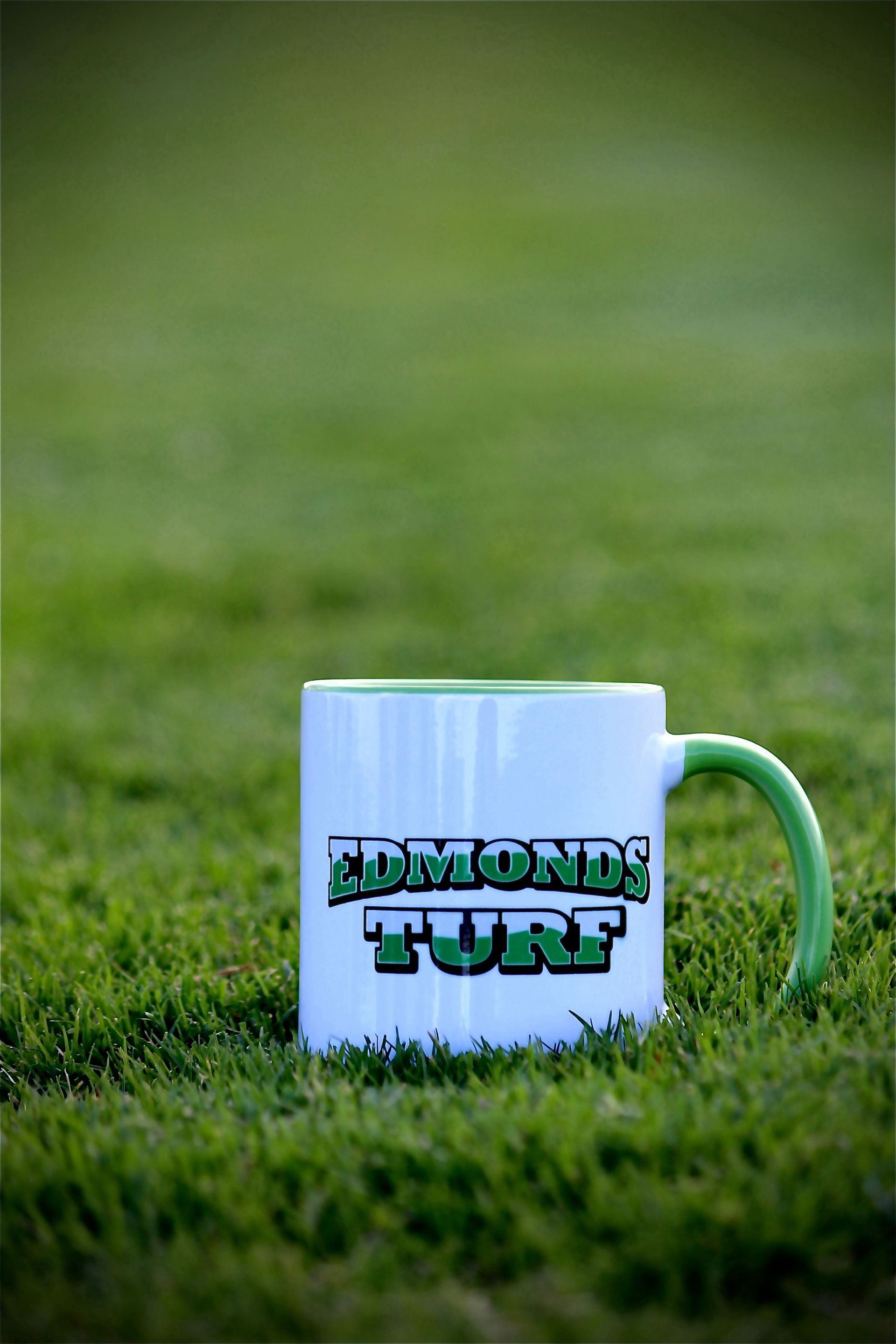 White mug with “Edmonds Turf” logo sitting on green grass outdoors