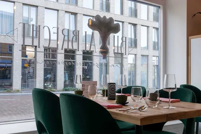 Een eettafel gedekt met wijnglazen en groene fluwelen stoelen voor een groot raam met restaurantreclame.