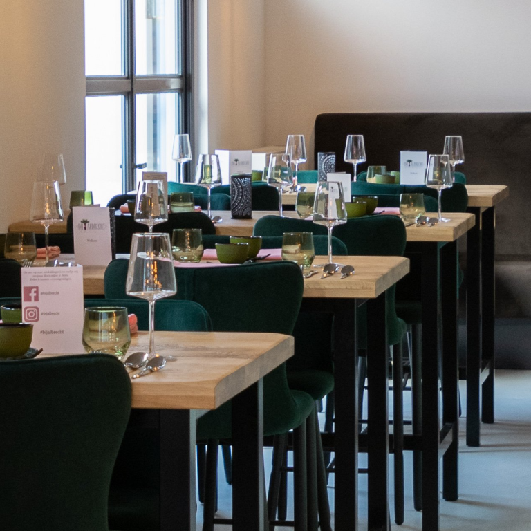 Een reeks houten eettafels met groene fluwelen stoelen en wijnglazen in een goed verlichte restaurantomgeving.