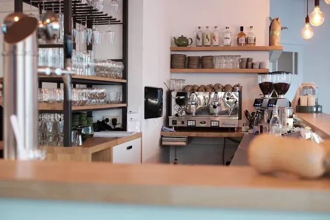 Een espressomachine en planken met glazen en flessen achter een houten toonbank in een lichte caféomgeving.