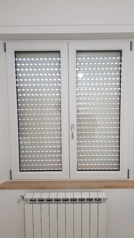 Finestra bianca con persiane chiuse e a lamelle; termosifone bianco in basso su una parete bianca.