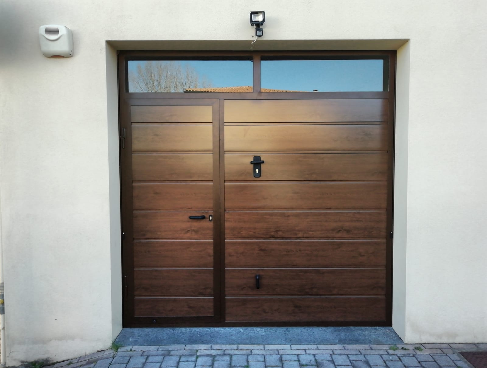 Porta da garage marrone con porta laterale, finestra e luce di sicurezza su parete color crema.