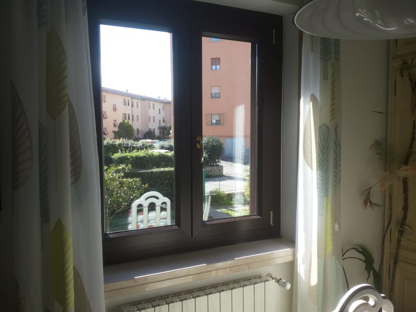 Finestra con vista su un giardino e su alcuni edifici in una giornata di sole.