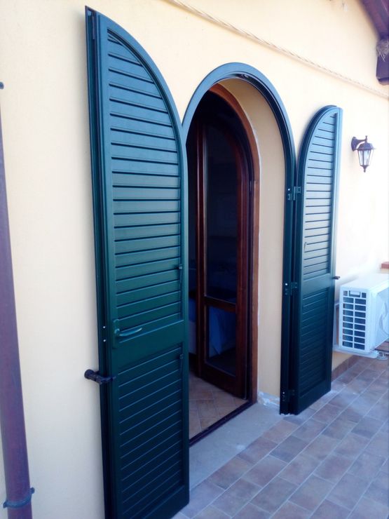 Persiane verdi ad arco si aprono su una porta di un edificio giallo chiaro.