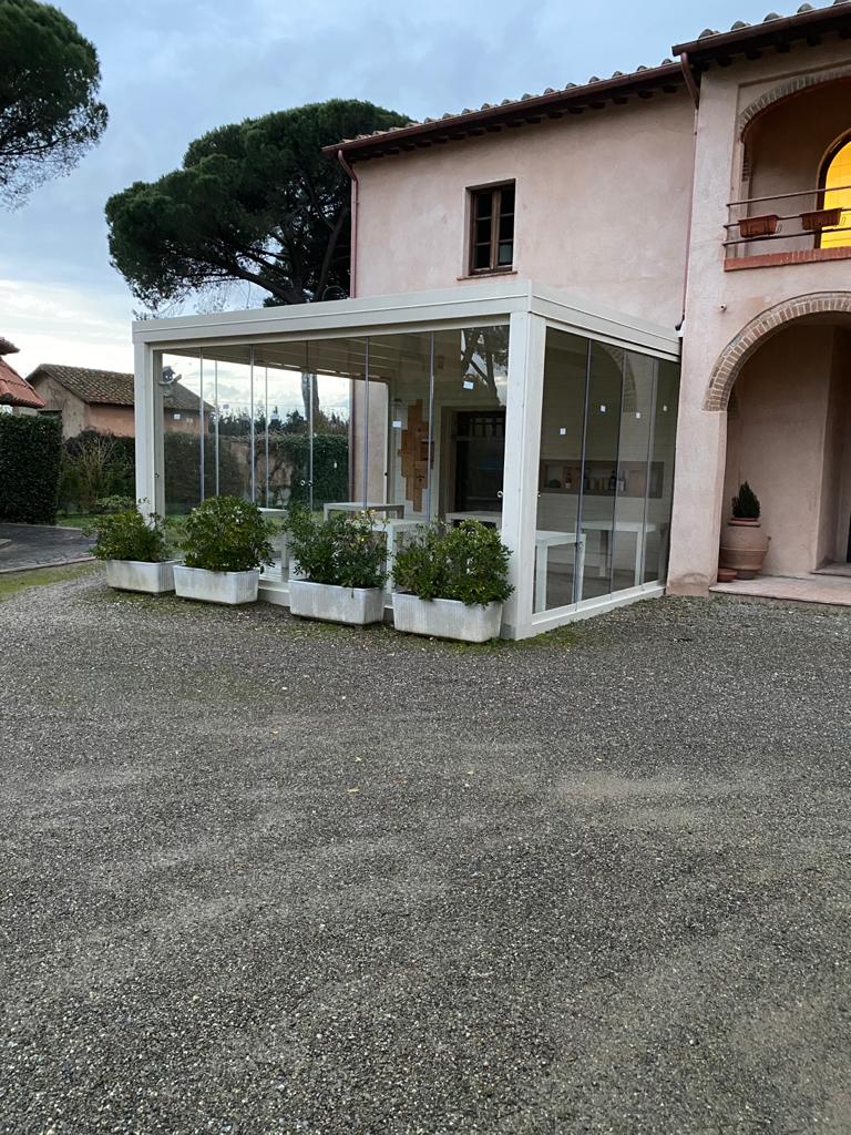 Un edificio beige con una zona pranzo esterna chiusa da vetrate. Fioriere bianche costeggiano le pareti in vetro; sentiero di ghiaia.