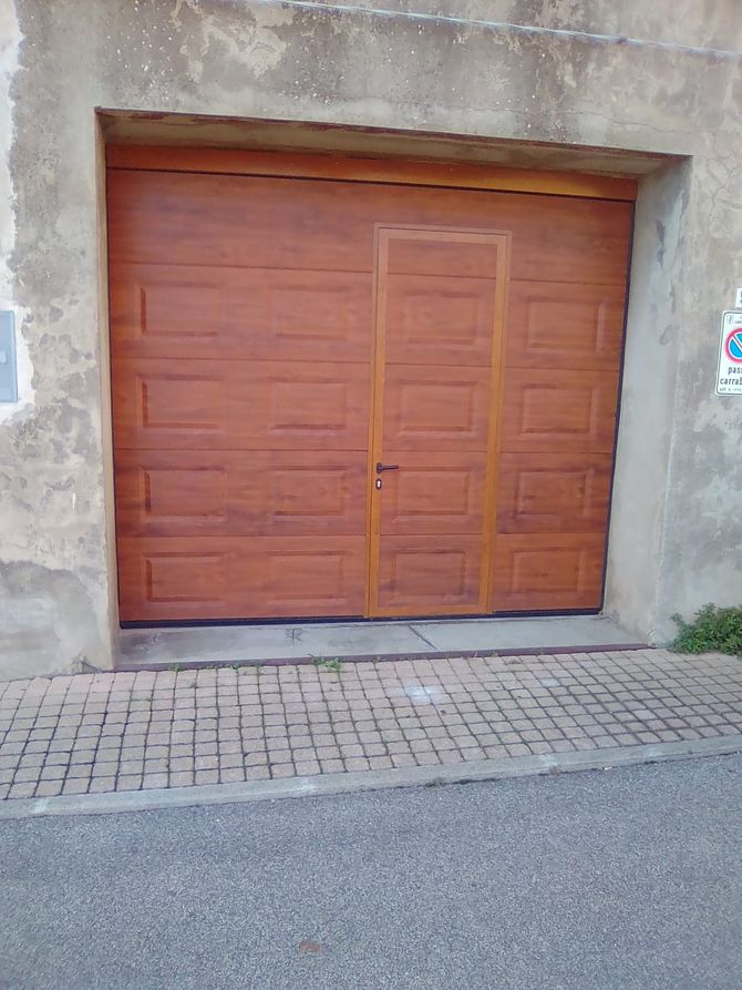 Porta da garage marrone con una porta più piccola inserita, incastonata in un muro di cemento invecchiato. Ciottoli e asfalto in primo piano.