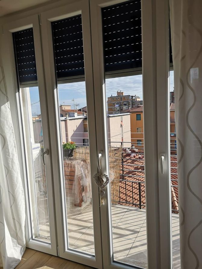 Interno di una casa moderna con finestre panoramiche che si affacciano su una città costiera al tramonto.