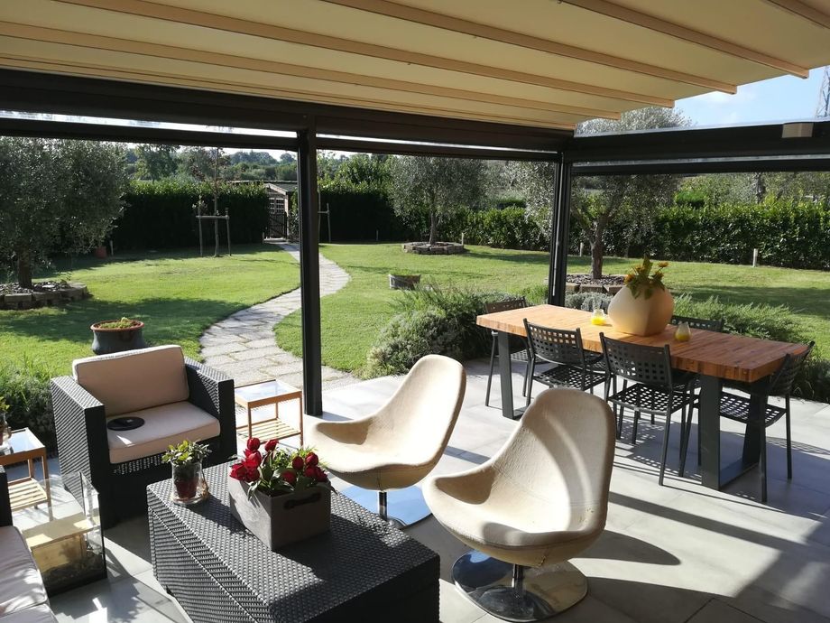 Pergola in legno con area salotto, barbecue e giardino lussureggiante nelle giornate di sole.