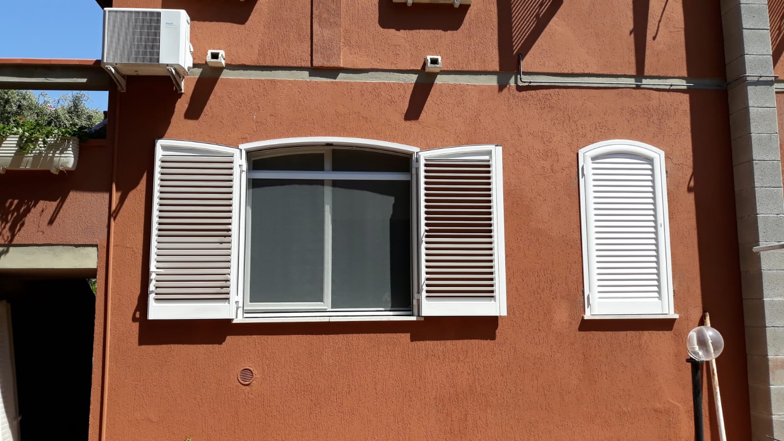 Edificio in stucco arancione con finestra e persiane bianche aperte a sinistra e chiuse a destra. Aria condizionata sopra.
