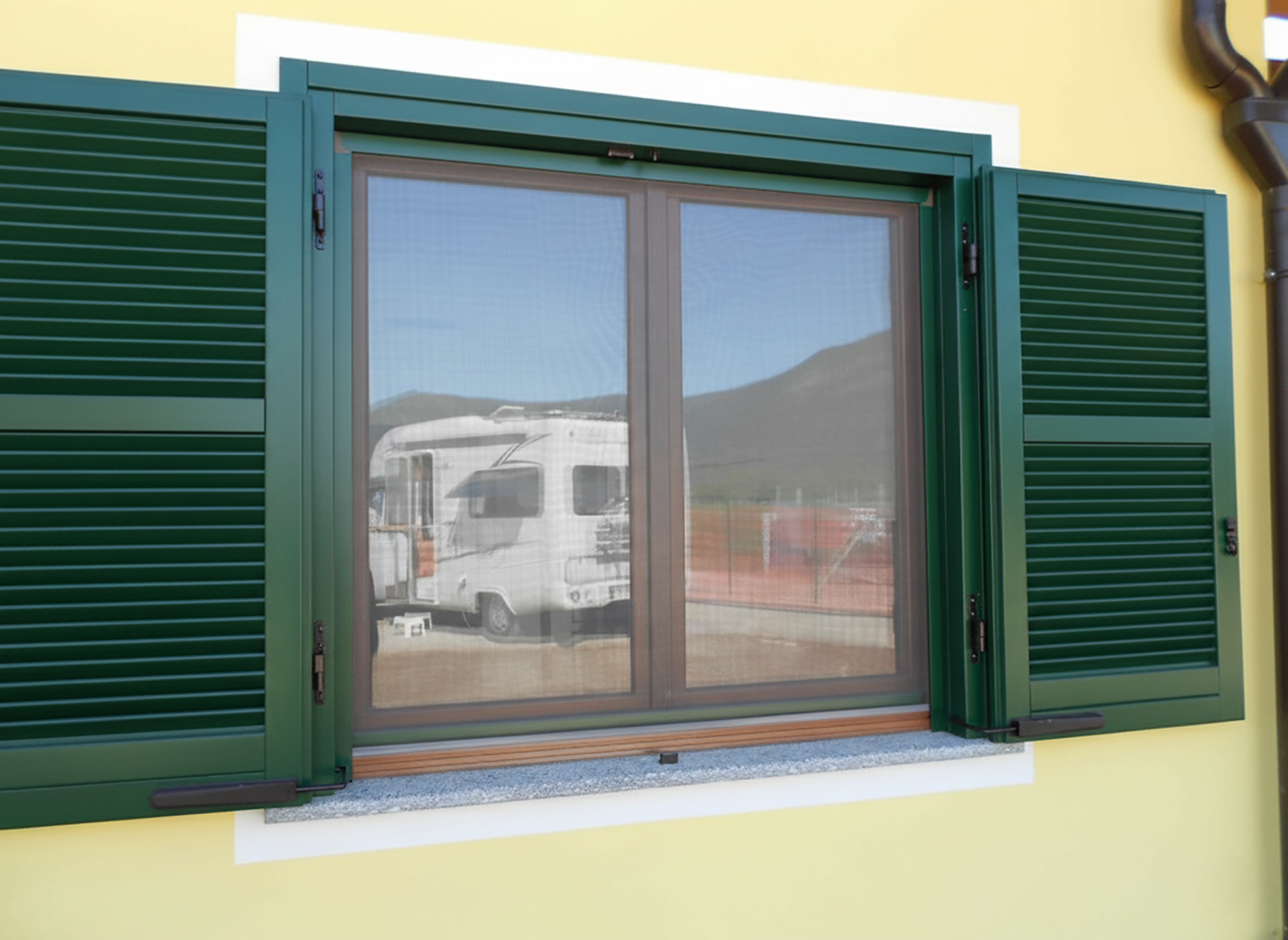 Le persiane verdi fiancheggiano una finestra con zanzariera; attraverso il vetro si vede un camper.