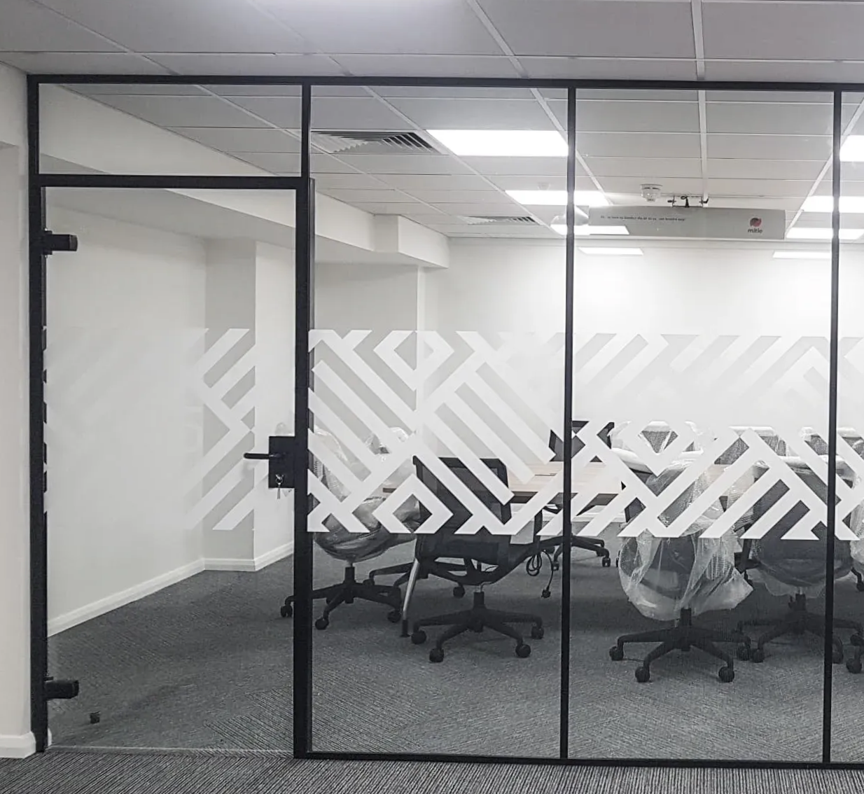 Glass-walled conference room with black frames, frosted design, and a black door. Chairs and projector are visible inside.