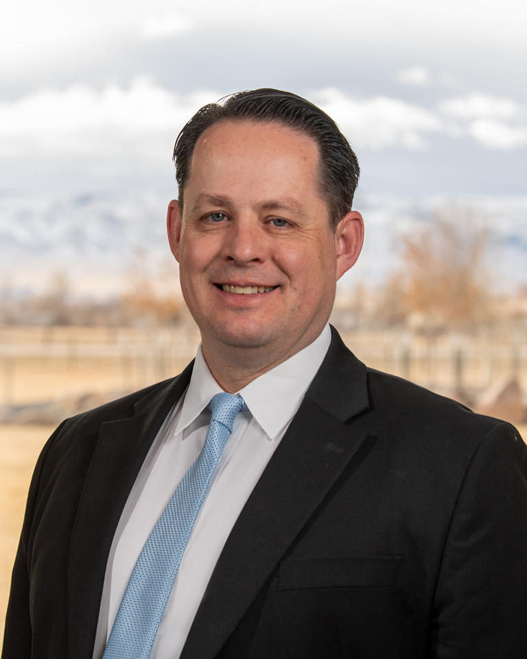Man in black suit and light blue tie smiles outdoors with mountains in background.