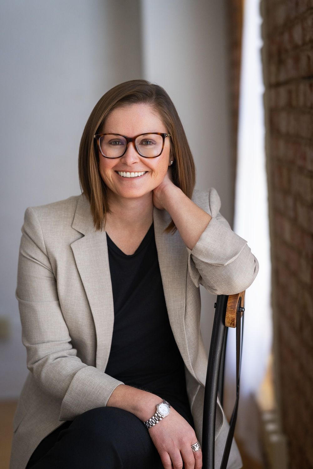 Woman in glasses and blazer smiles, sitting on a chair, hand on neck. Brick wall in background.