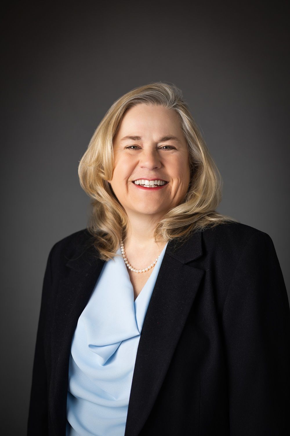 Woman in a black blazer and light blue blouse smiling, wearing a pearl necklace against a gray backdrop.
