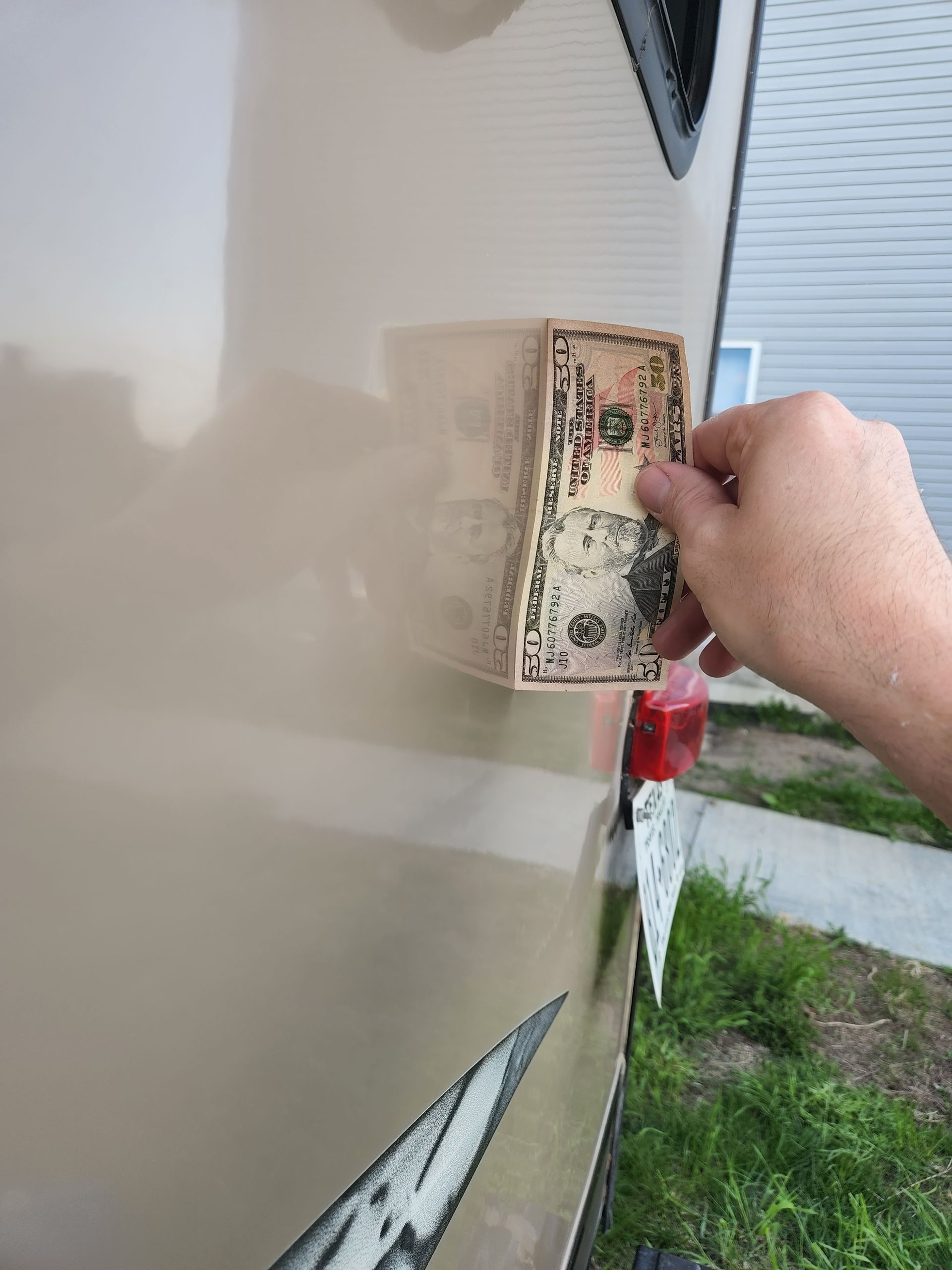 Hand holding a bill against the side of a beige vehicle outdoors near grass and a driveway