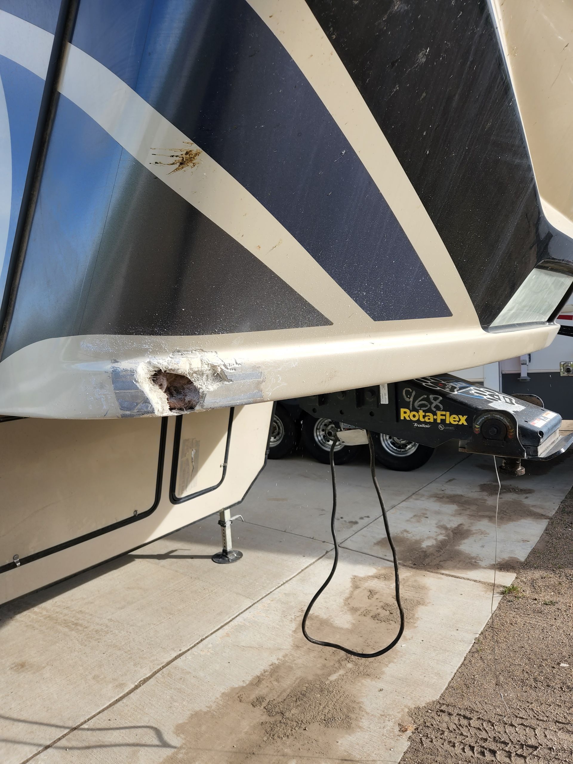 Damaged camper trailer corner with a hanging cable and dark stain on the concrete driveway