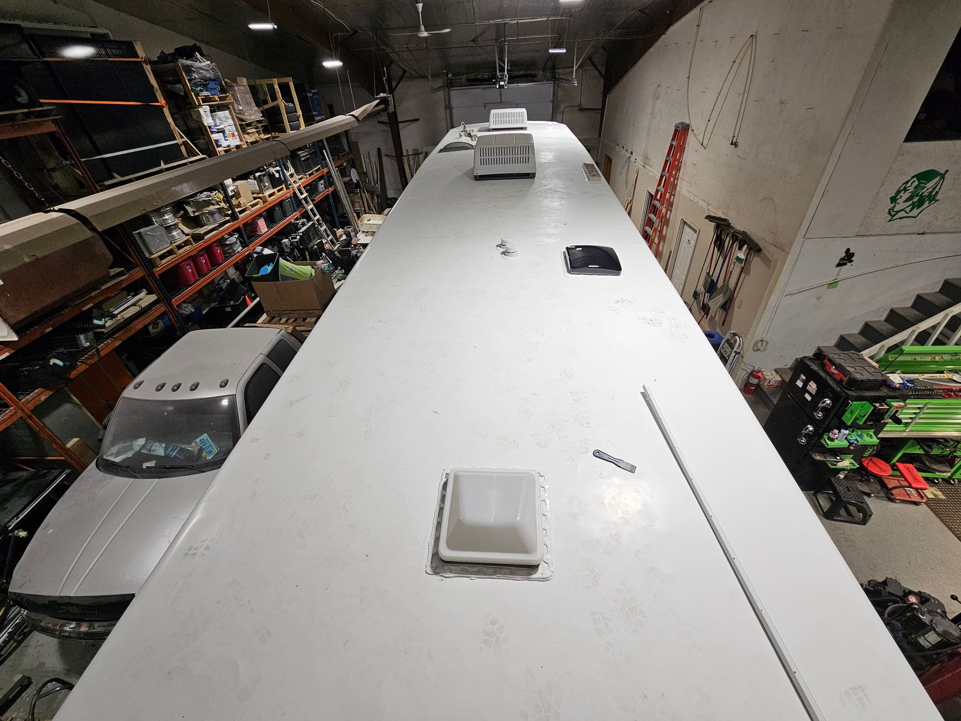 Inside a warehouse, a large white vehicle roof with vents is viewed from above, surrounded by shelving and equipment.