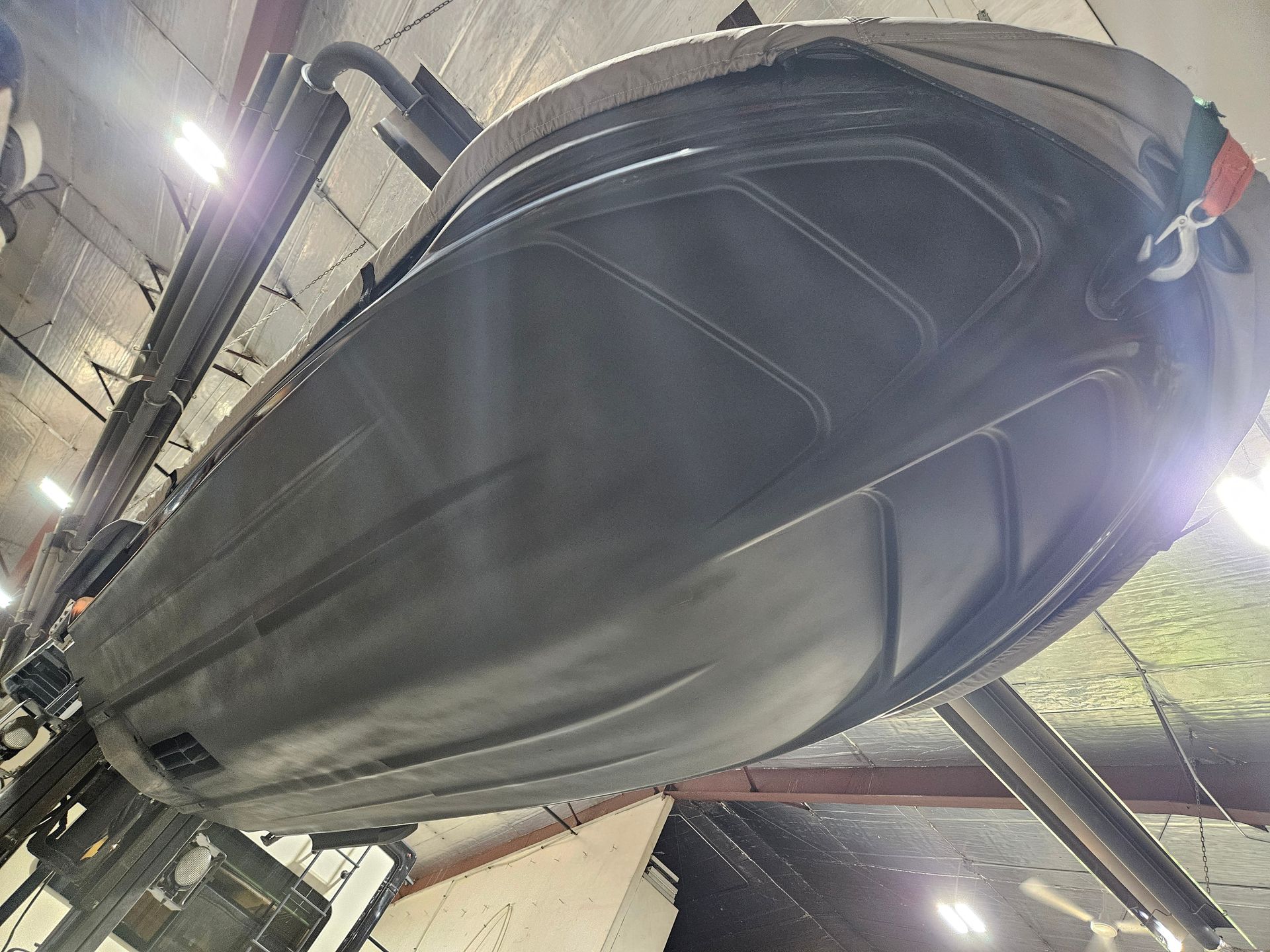 Large black boat hull being worked on in a shipyard, viewed from below against overhead lights.