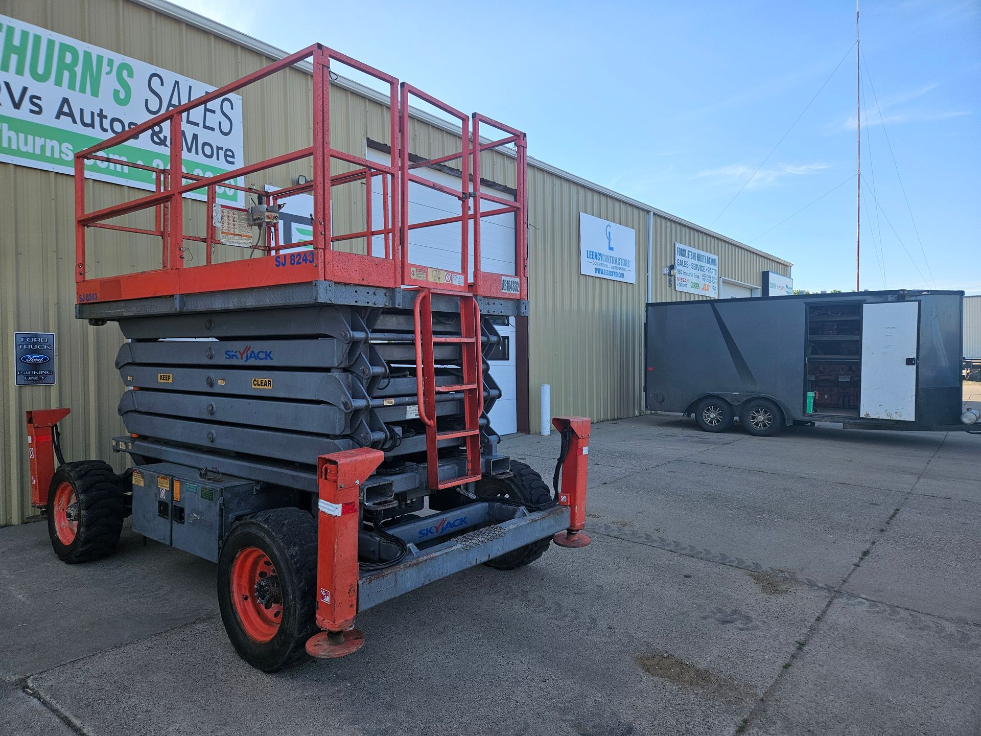 Red scissor lift with orange guardrails parked outside a warehouse on a sunny day.