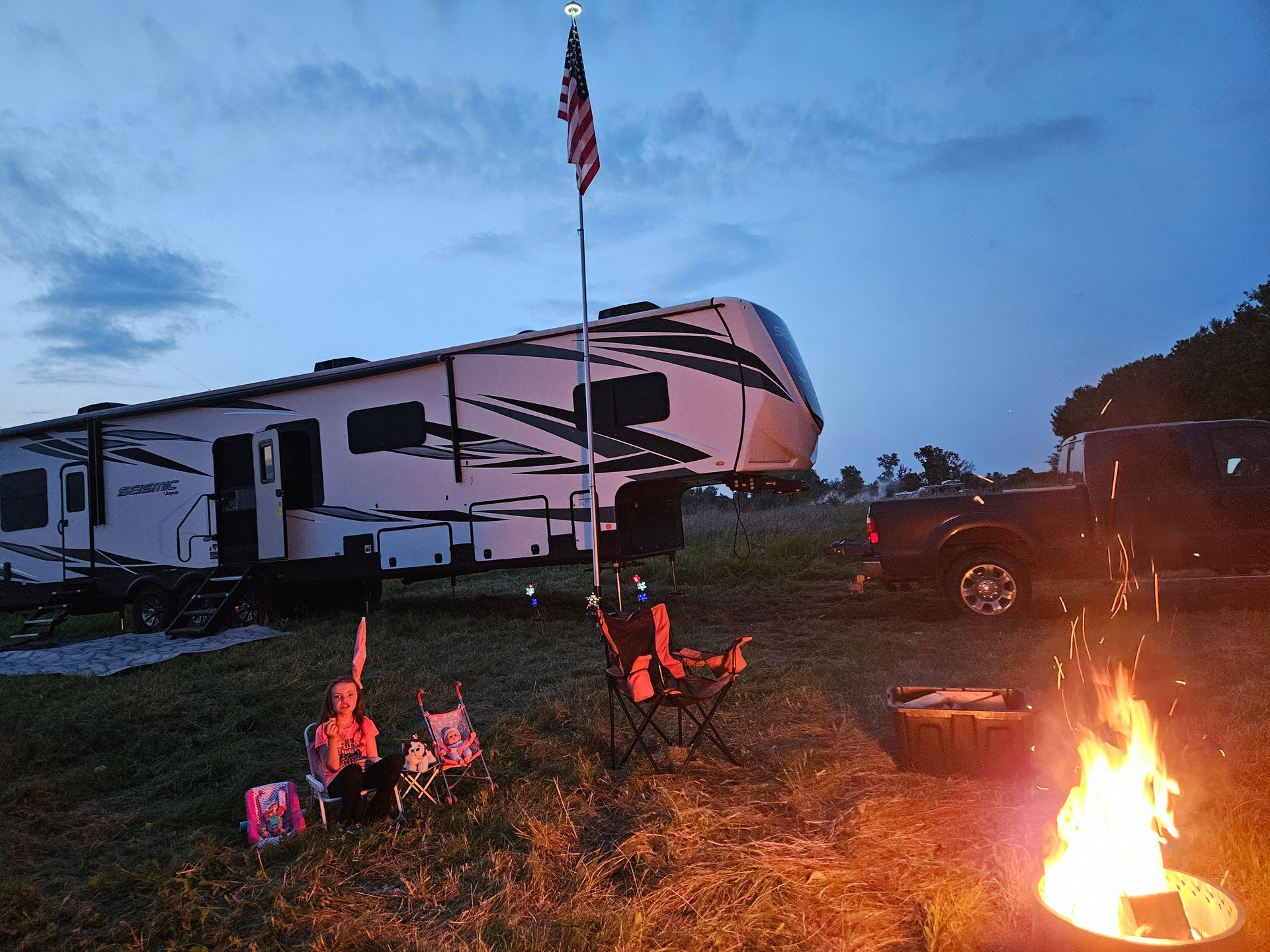 RV campsite at dusk with a campfire, lawn chairs, and an American flag