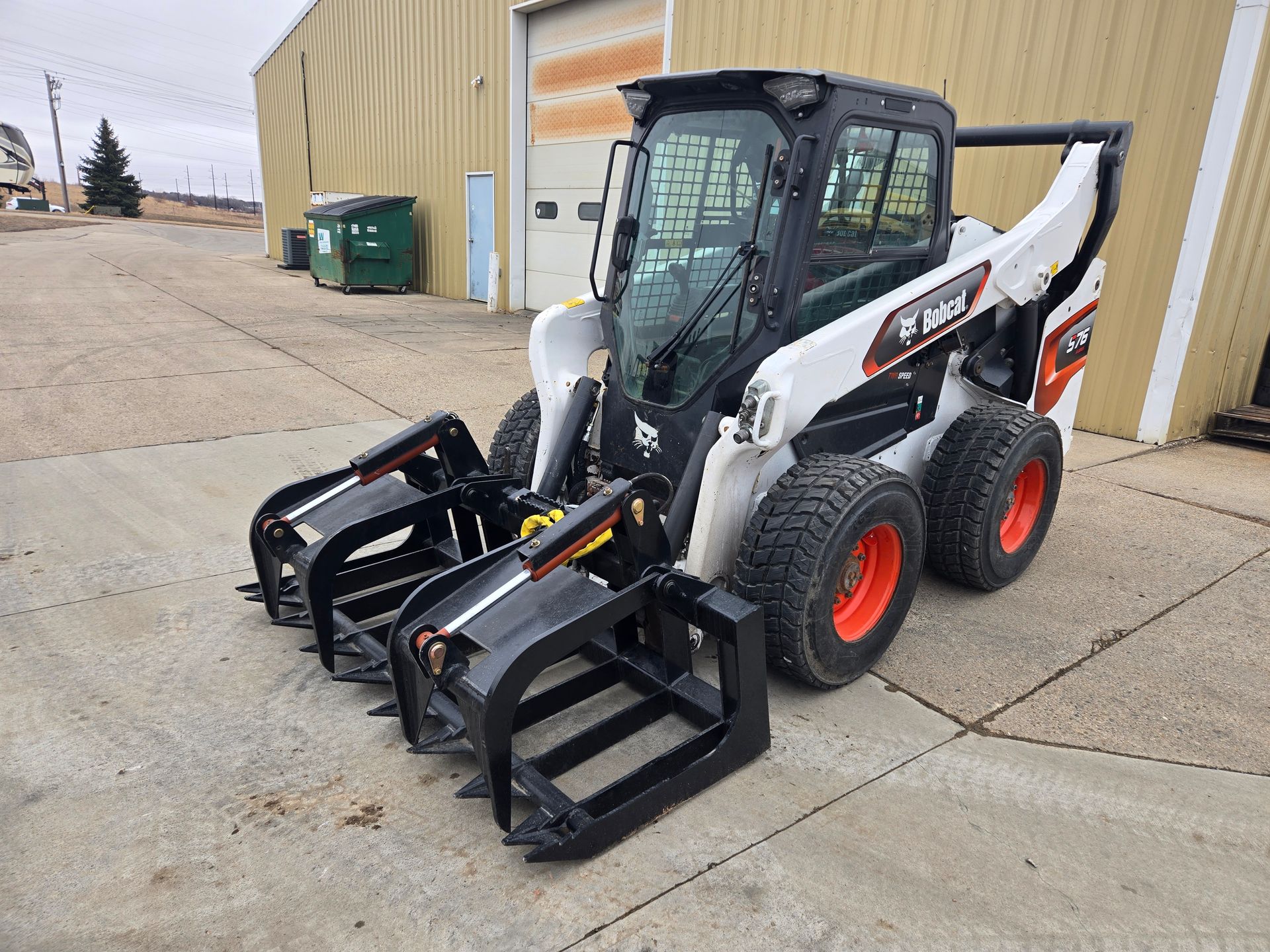 White skid steer loader with dual front grapples parked outside a beige building