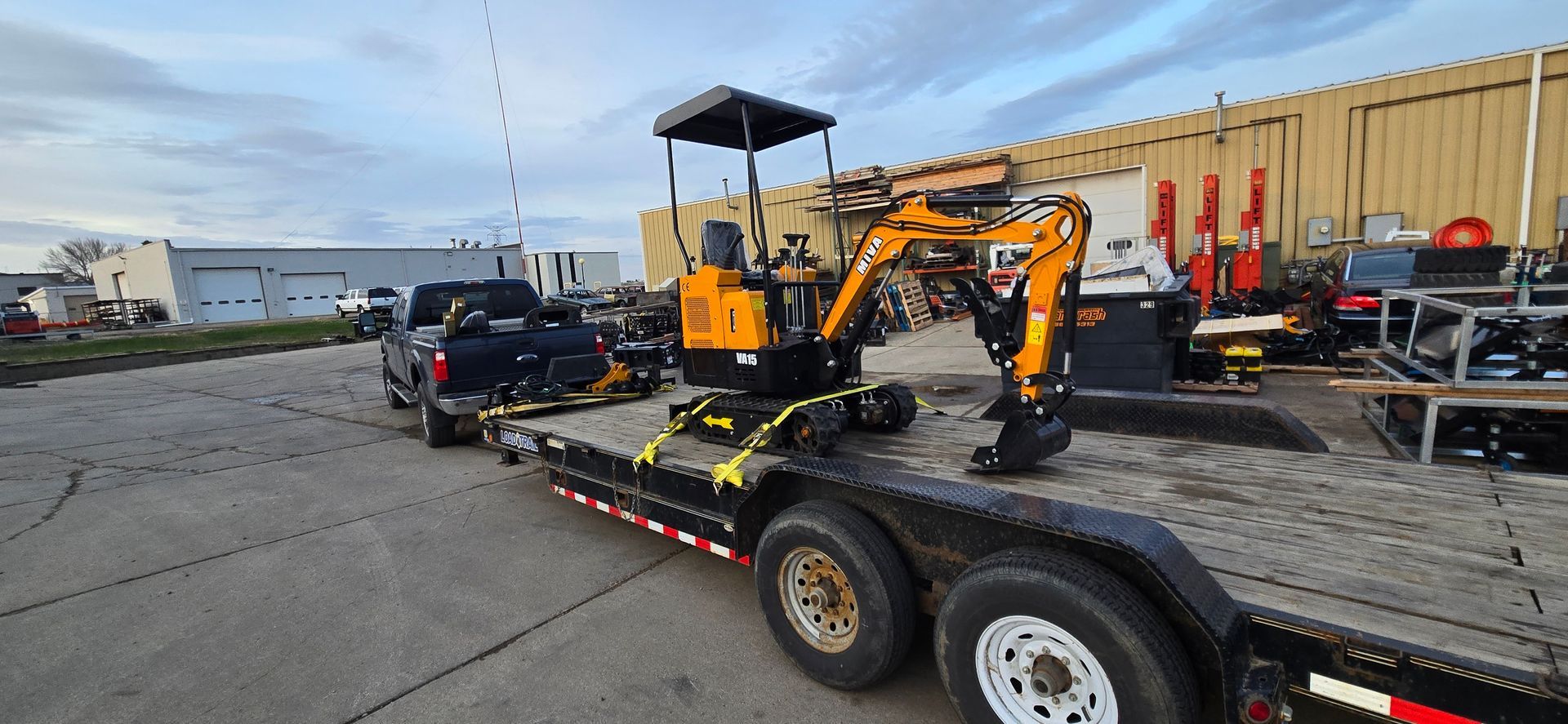 Orange mini excavator on a trailer in an industrial lot, with trucks and equipment in the background