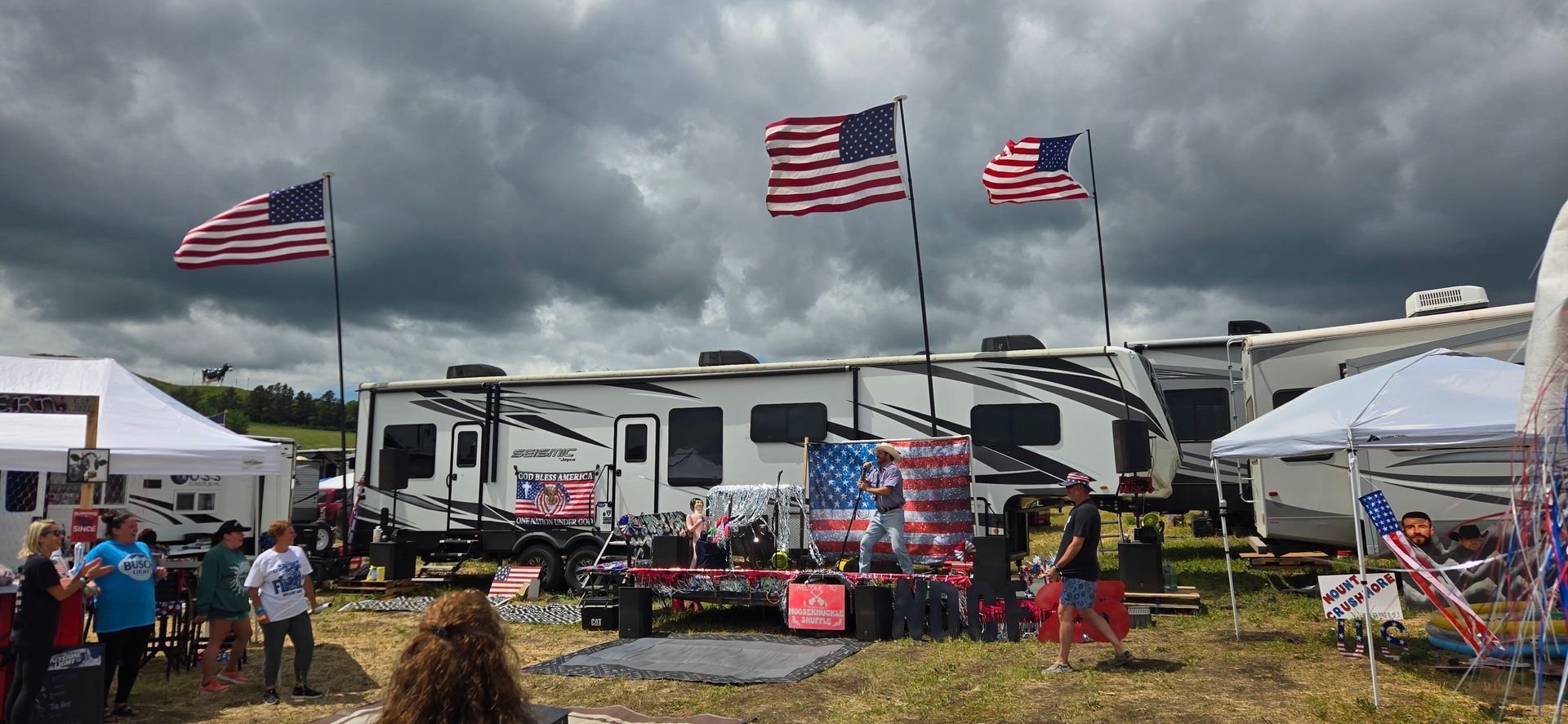 Outdoor RV event with American flags, tents, and people gathered under cloudy skies.