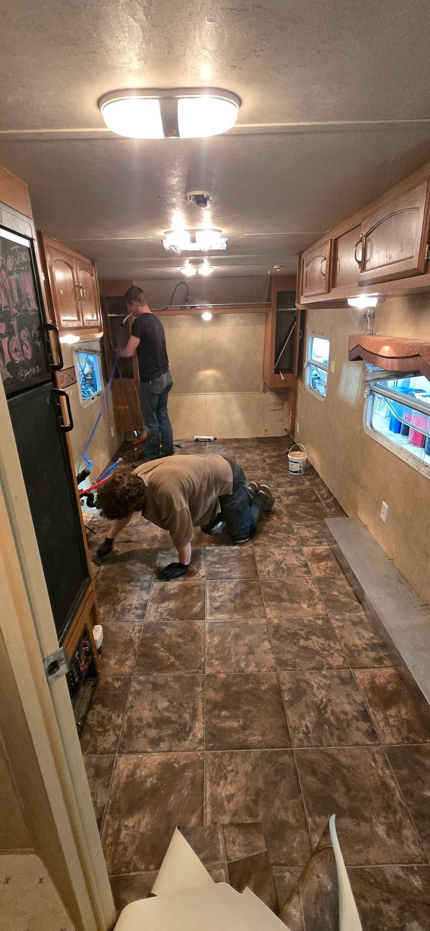 Inside a narrow RV kitchen, two workers install or inspect cabinetry and flooring.