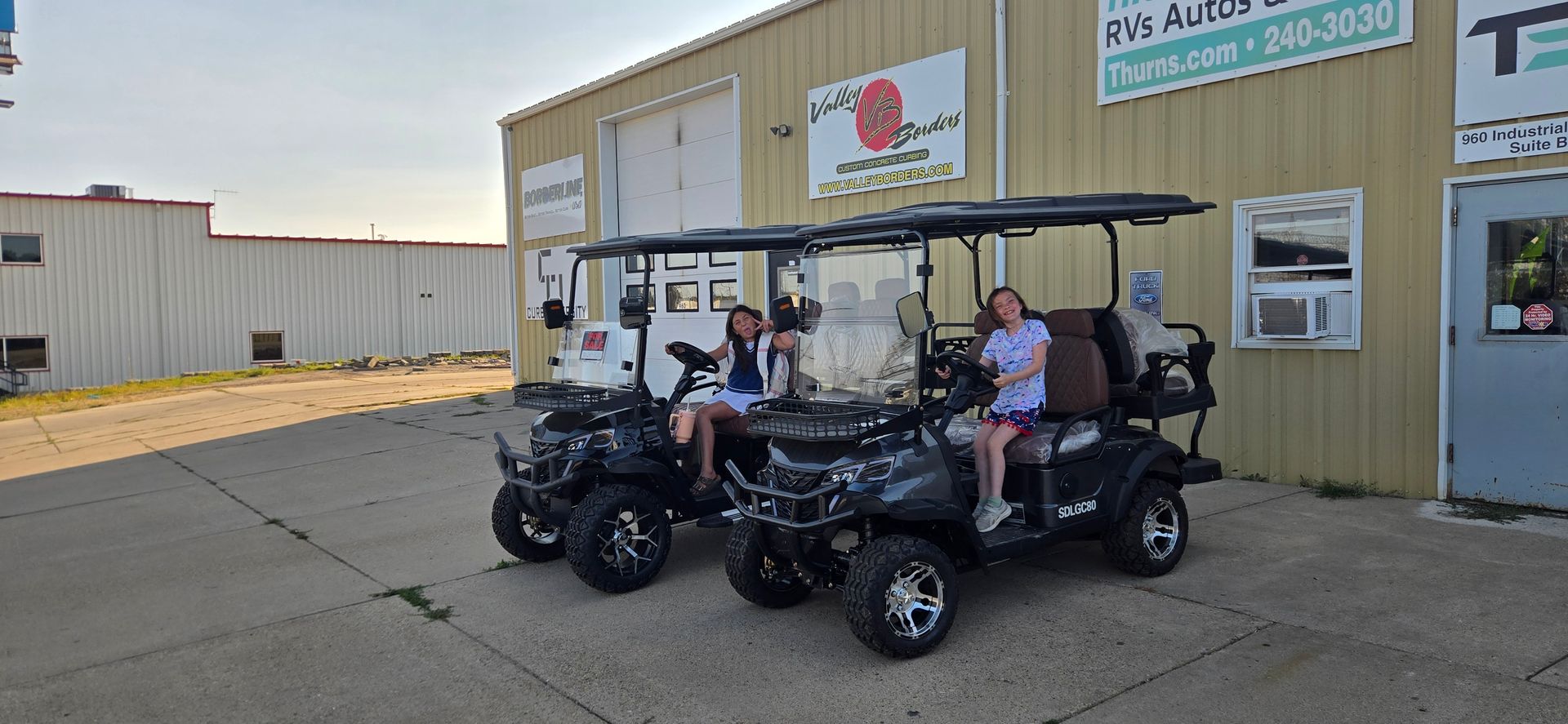 Two people on a black utility cart outside an industrial building with a white sign and orange logo.