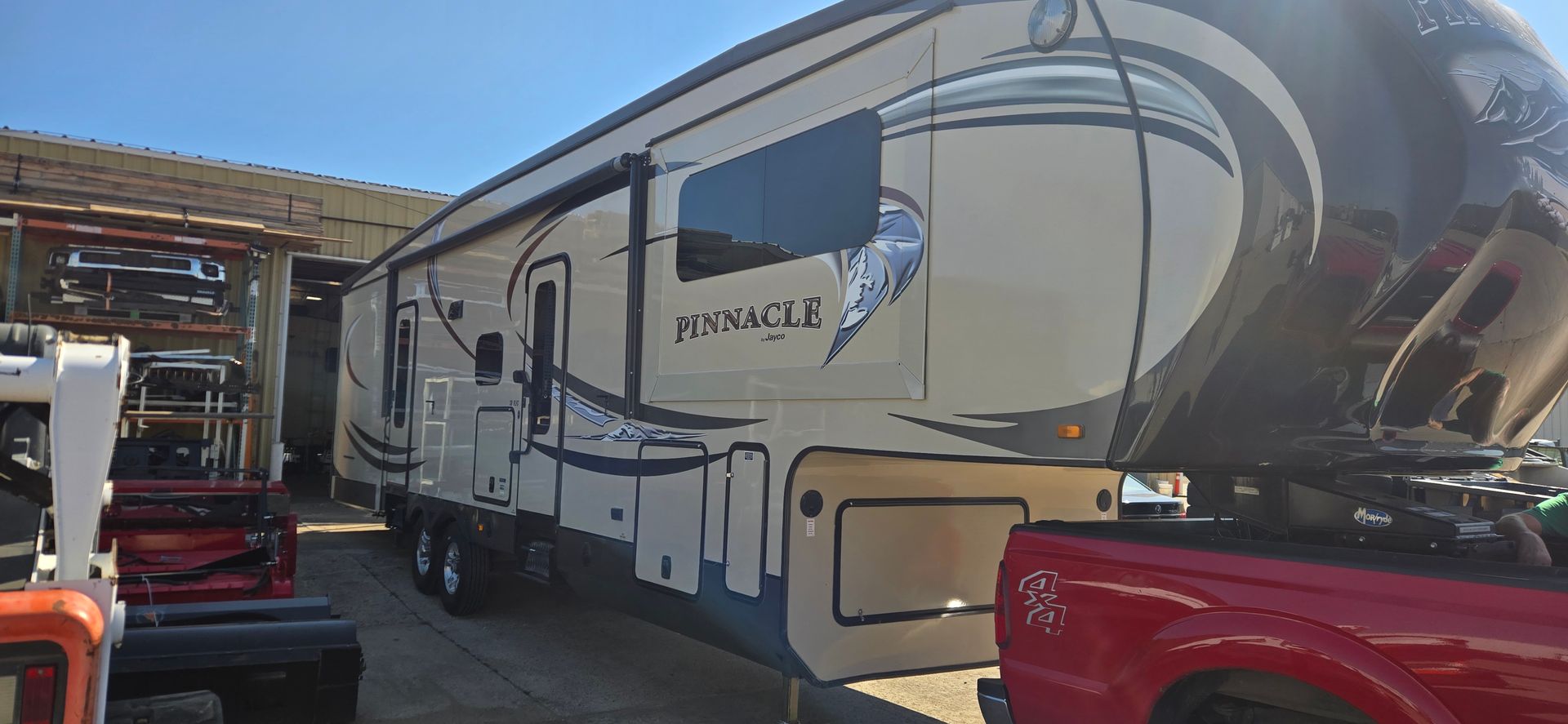 Large beige and brown RV parked between a red truck and a metal carport in a lot