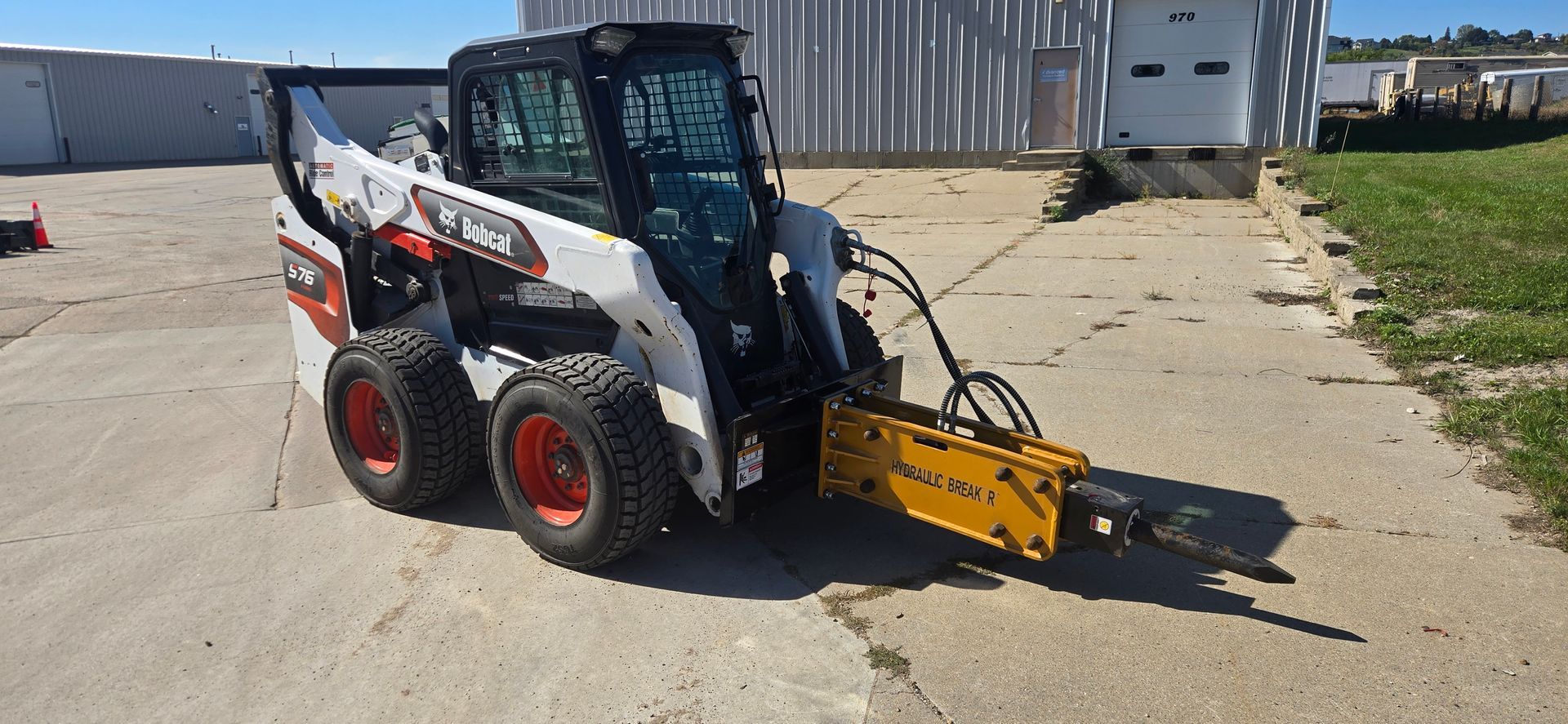 Compact skid steer with a yellow attachment on a gravel lot near a building and grass.