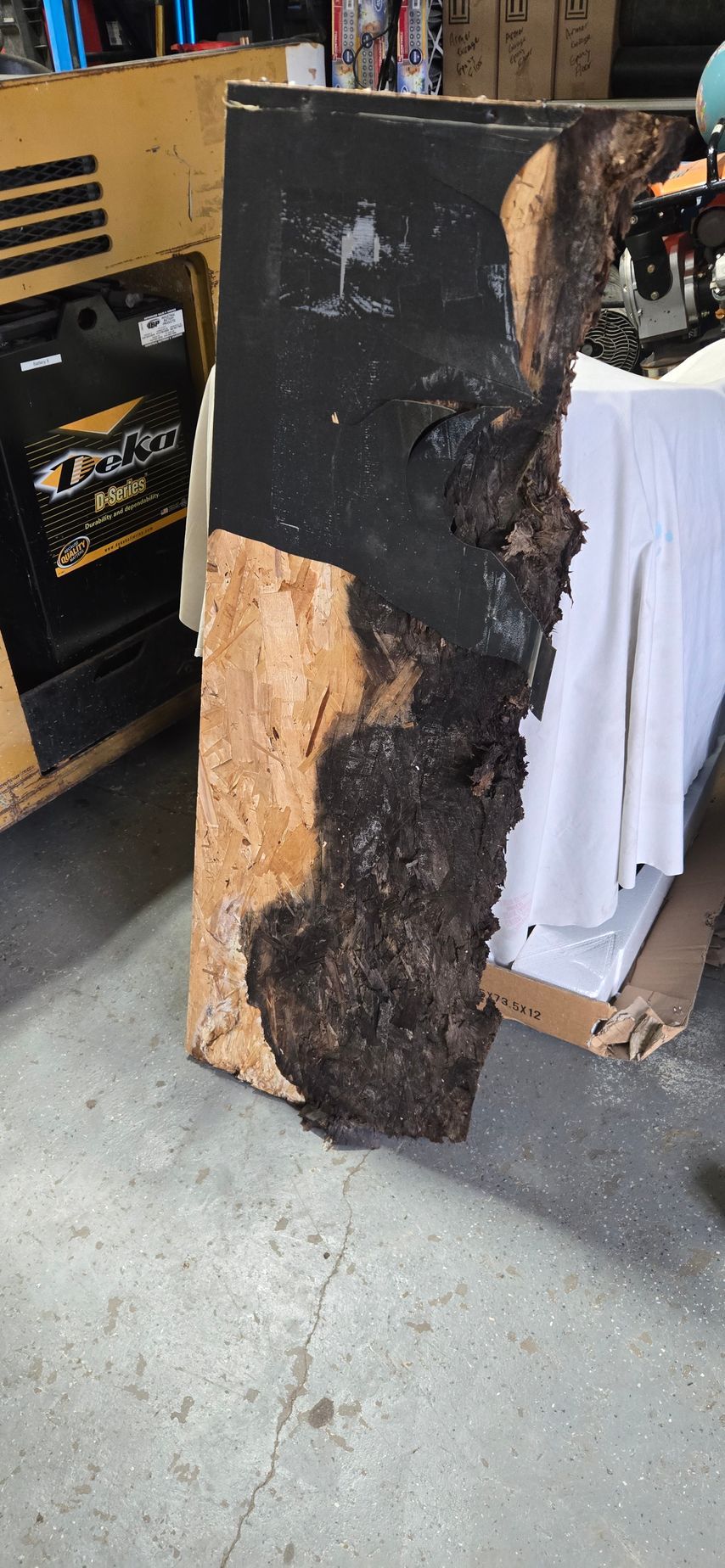 Charred wooden board with a large burned section, leaning in a workshop or garage.