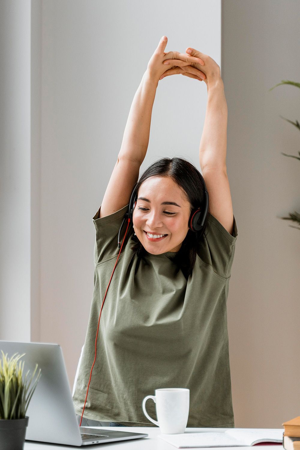 Woman in green shirt stretches arms overhead, smiling, at a desk with laptop, headphones, and coffee cup.