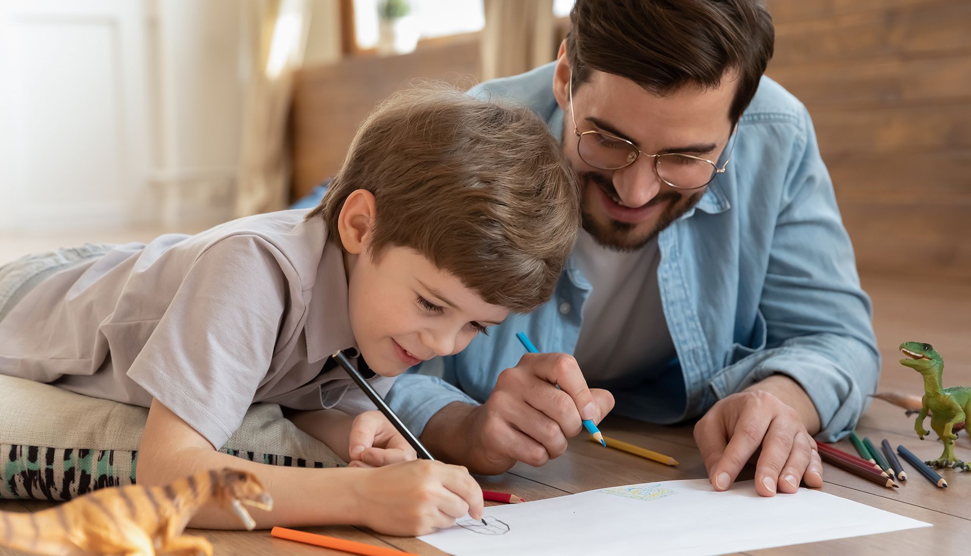 Boy and man drawing together on floor; both smiling and focused, with toys nearby.