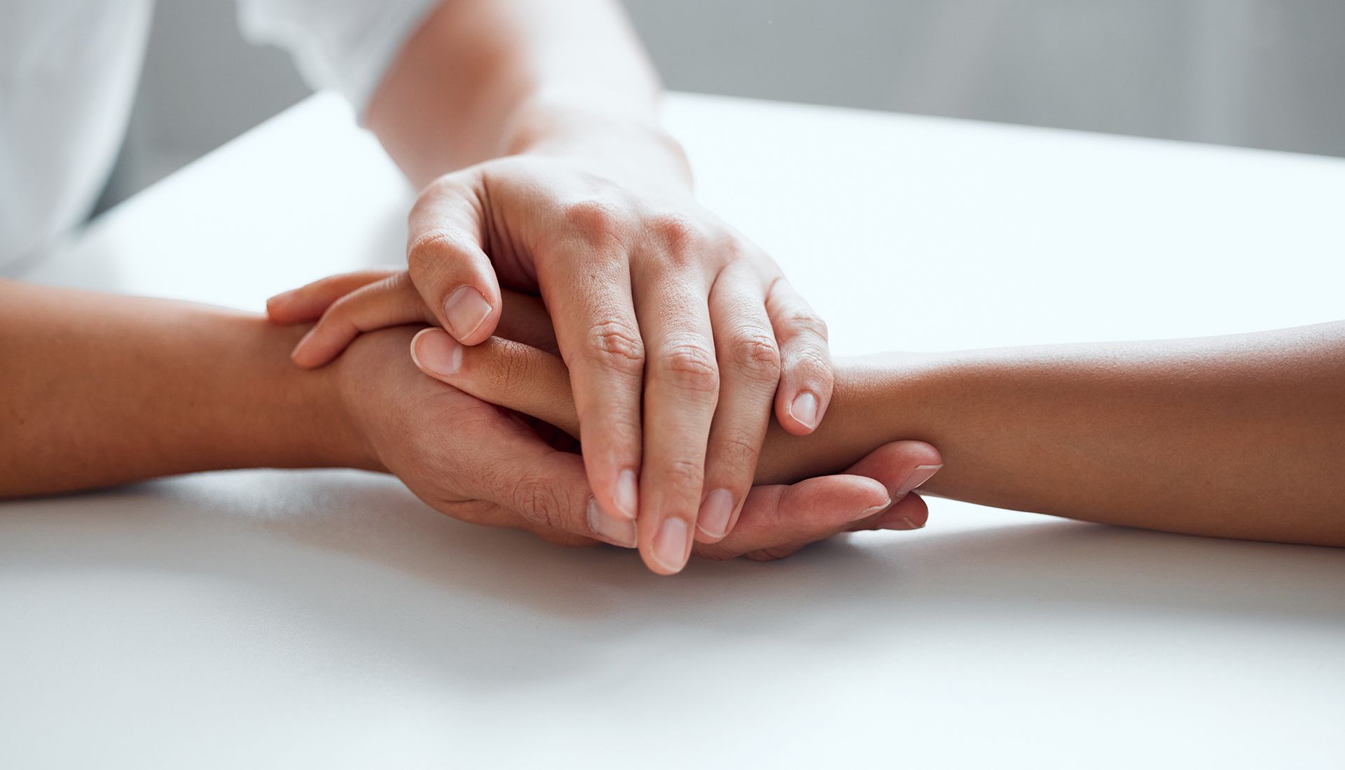 Hands clasped on a white surface; one person's hand over another, offering support and comfort.