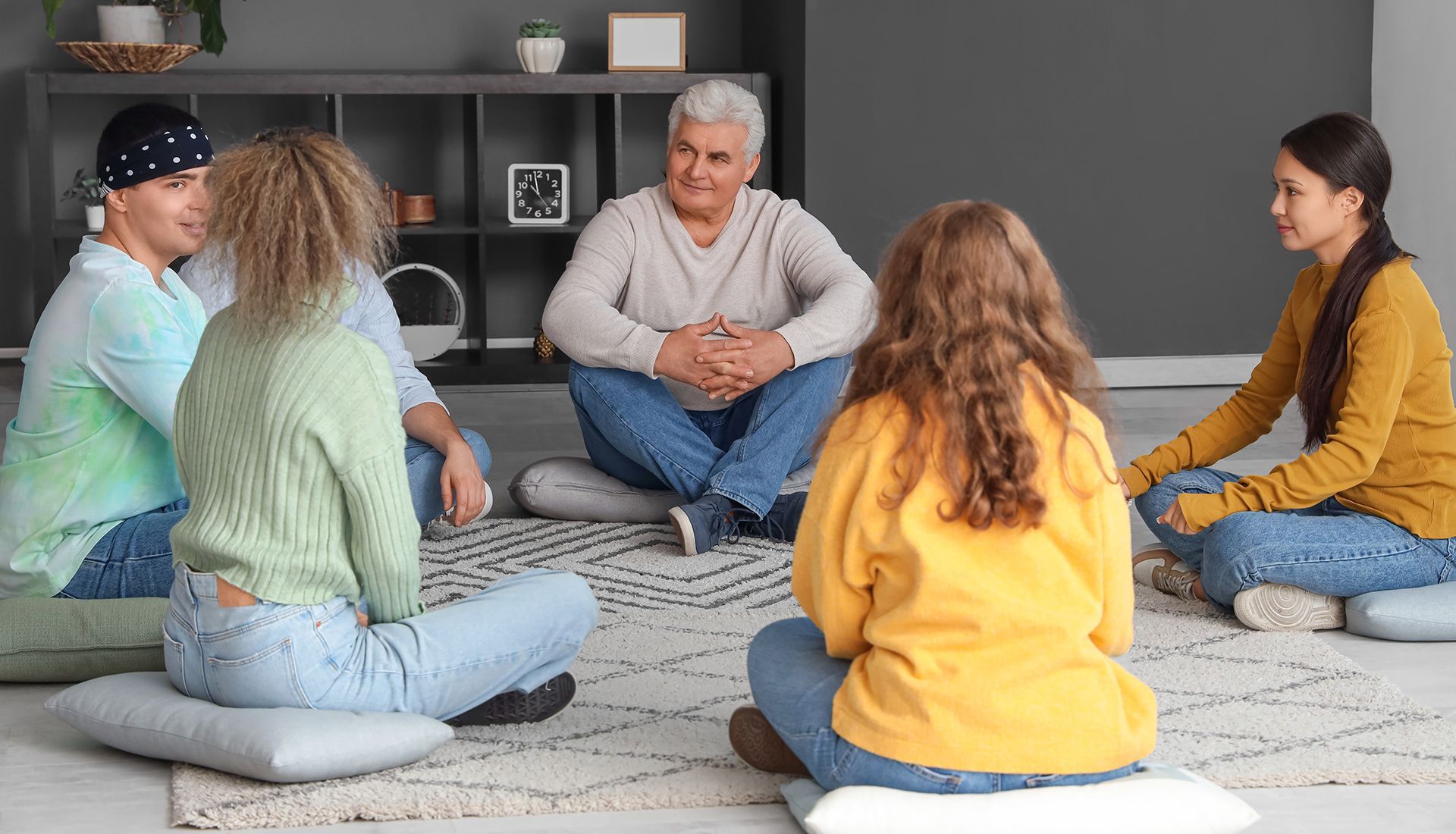 People in a circle on cushions, listening to an older man. Indoor setting, likely a therapy or support group.