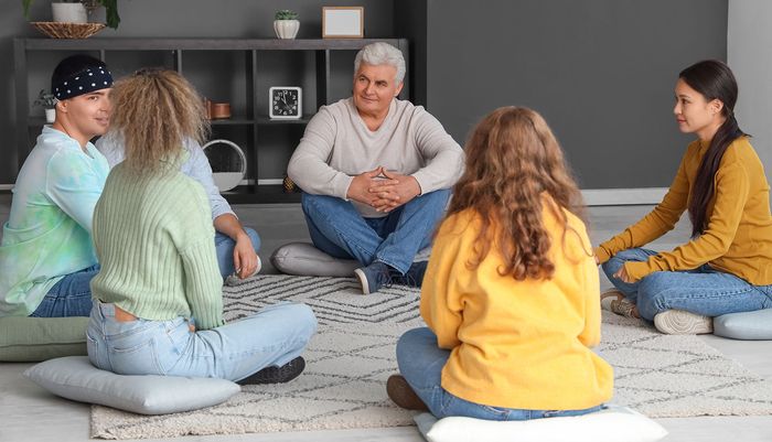 People in a circle on cushions, listening to an older man. Indoor setting, likely a therapy or support group.
