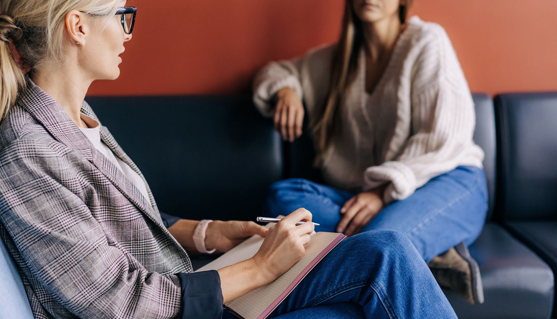 A therapist in glasses taking notes; client in jeans sits on a couch.