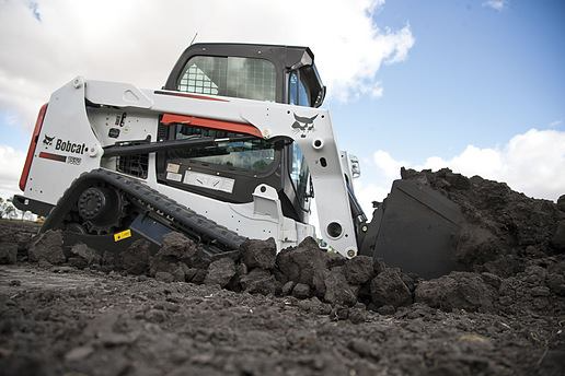 White Bobcat compact track loader digging into dark soil under a cloudy sky.