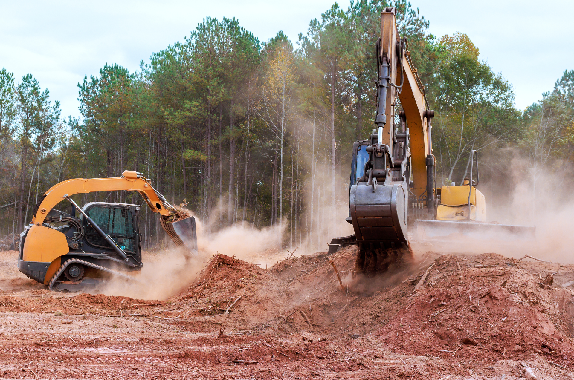 Two excavators, yellow and orange, digging up red dirt, creating dust clouds, in a wooded area.