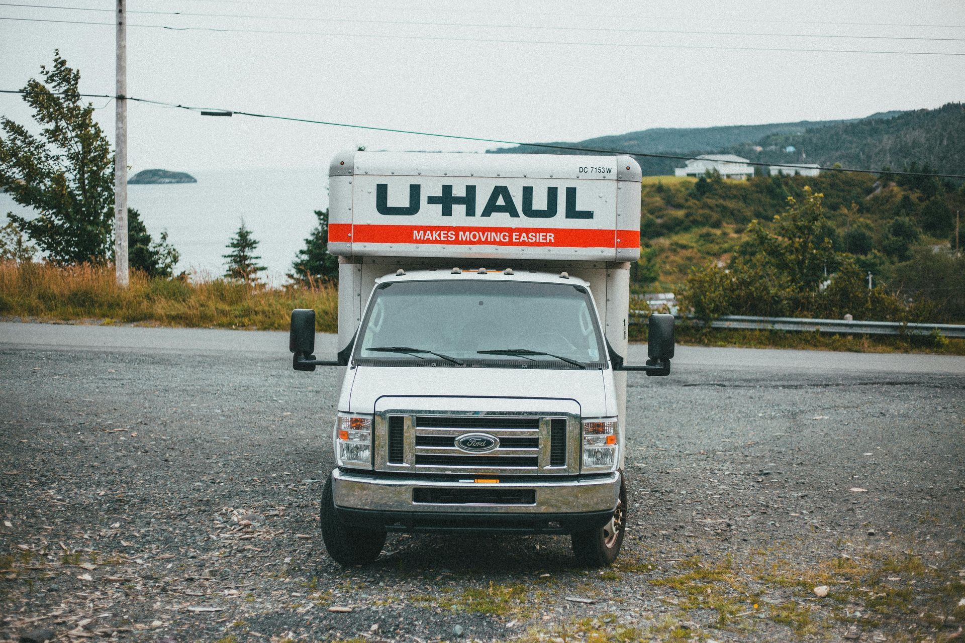 White U-Haul truck parked on gravel, facing forward. Ocean and hills in the background on an overcast day.