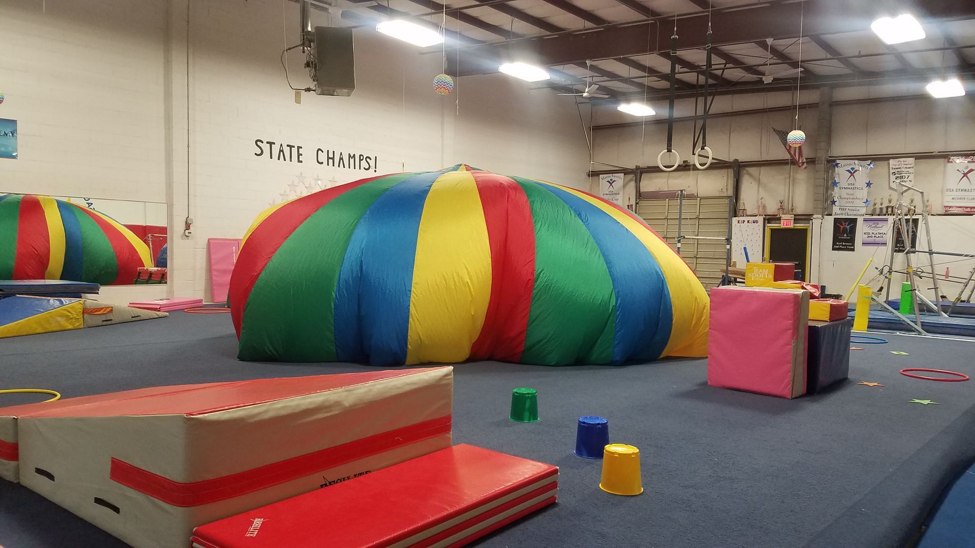 A colorful parachute dome on a gymnastics floor with various training mats and obstacles.