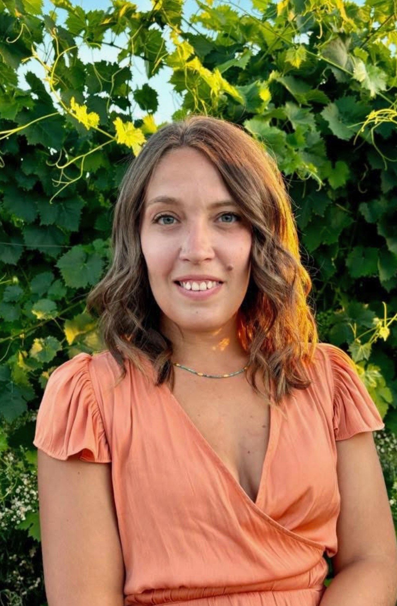 A person with shoulder-length wavy hair, wearing a peach-colored dress, smiling in front of a lush green vine background.