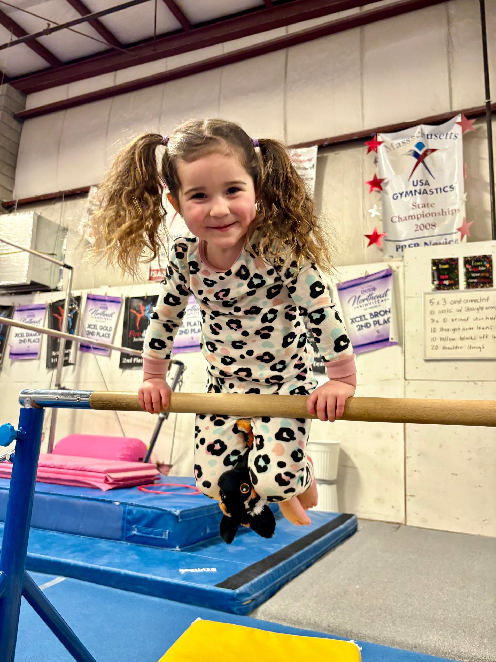 A smiling child with curly hair in pigtails wearing leopard-print pajamas, balancing on a wooden gymnastic bar.