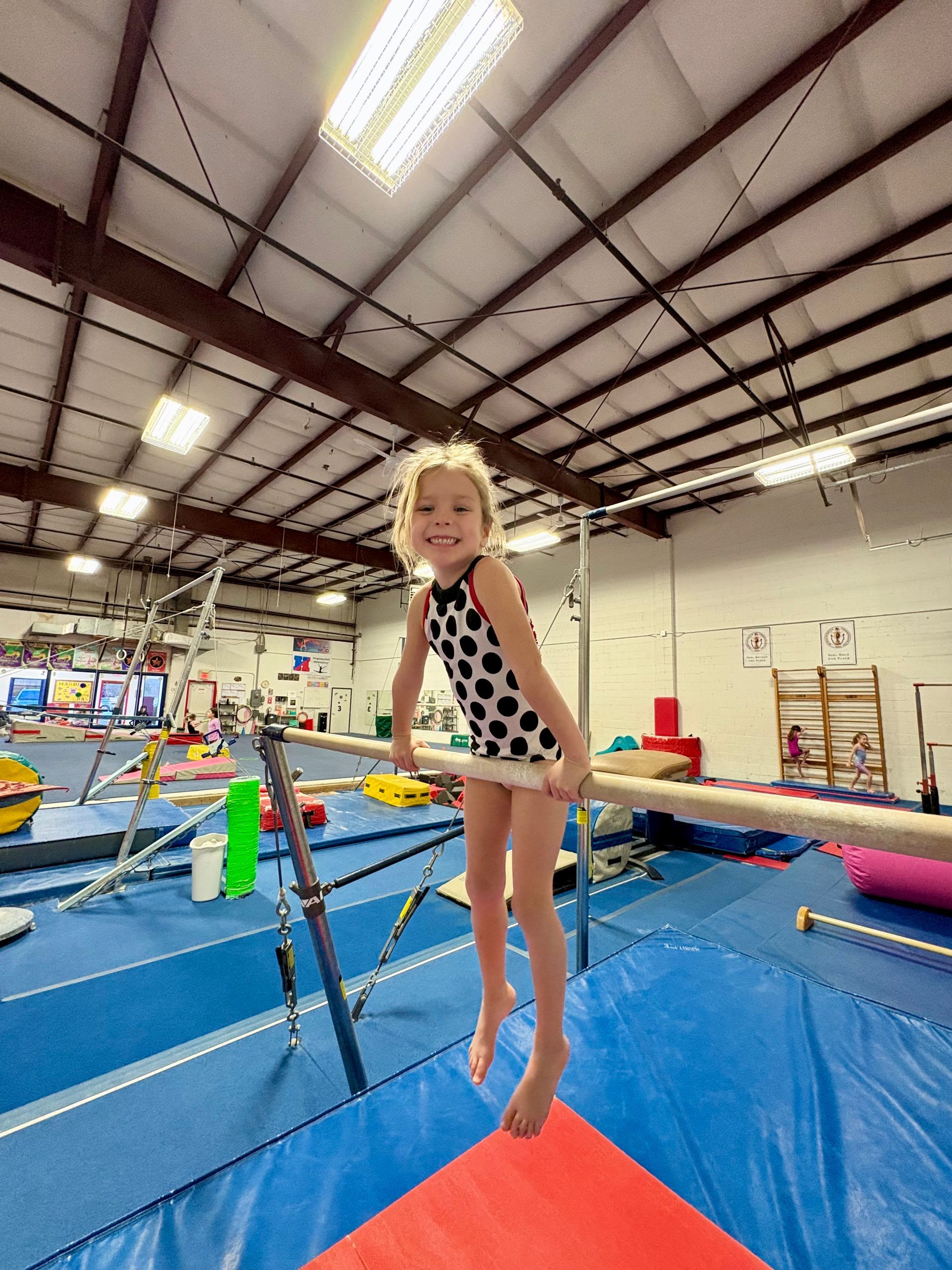 A young gymnast in a polka-dot leotard smiling while holding onto the uneven bars in a gym.